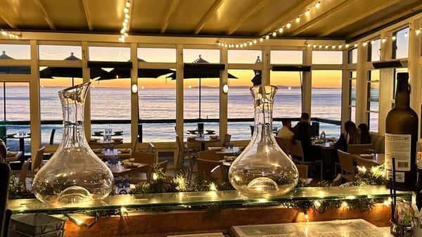Romantic oceanfront dining scene at Driftwood Kitchen in Laguna Beach, featuring elegant glass wine decanters, string lights, and a golden sunset over the Pacific Ocean viewed through large windows.