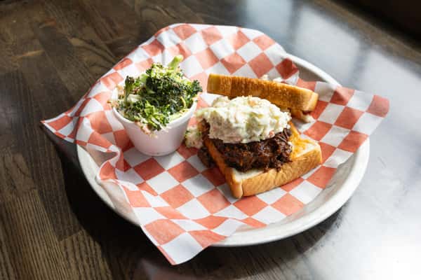Open-faced pulled pork sandwich with coleslaw and toasted bun alongside a fresh broccoli salad side