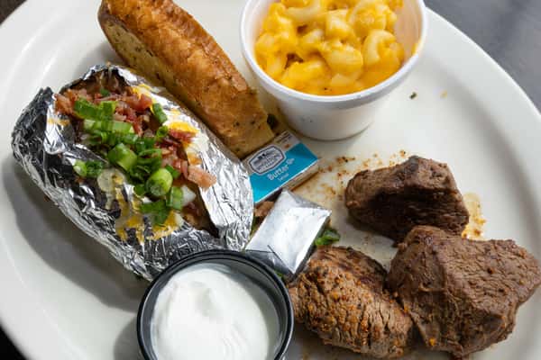 Grilled steak tips with a loaded baked potato, mac and cheese, garlic toast, and sour cream on a white plate
