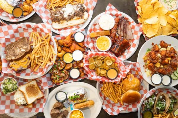 Overhead spread of bar food including BBQ ribs, brisket, wings, burgers, fries, fried pickles, and salads on checkered paper-lined plates