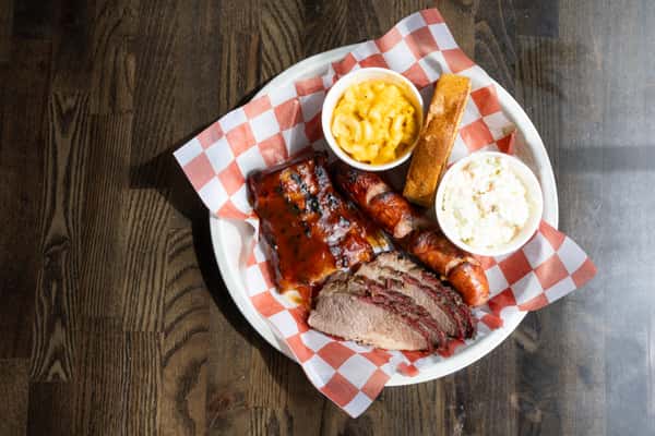 BBQ sampler with glazed ribs, smoked sausage, sliced brisket, mac and cheese, coleslaw, and cornbread on a checkered plate