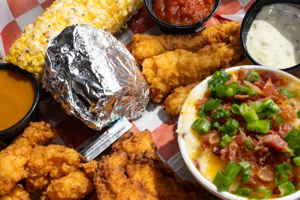 A close-up, top-down view of a hearty seafood platter served on red and white checkered paper. The meal includes fried shrimp, fried fish fillets, a cob of corn with butter, and a foil-wrapped side. There is also a bowl of creamy grits or mashed potatoes topped with melted cheese, bacon bits, and sliced green onions. Three small dipping cups containing cocktail sauce, tartar sauce, and a golden gravy or sauce are positioned around the tray.