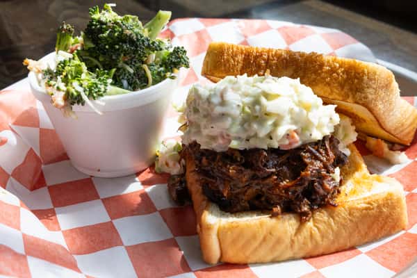 Pulled pork sandwich topped with creamy coleslaw on a toasted bun, served with a side of broccoli salad