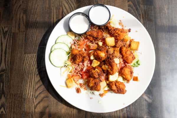 Overhead view of buffalo chicken salad with crispy fried chicken, bacon, tomatoes, cucumbers, shredded cheese, and ranch dressing on a white plate
