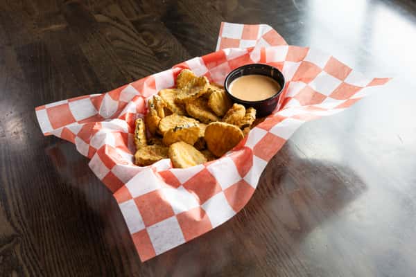 Crispy fried pickle chips served with a creamy dipping sauce in a red checkered basket on a wooden table