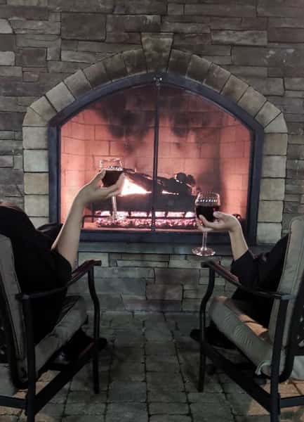 Two people sitting in chairs holding wine infront of a fireplace