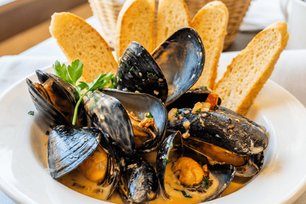 Steamed mussels in a cream sauce with visible herbs, served with baguette slices and a parsley garnish.