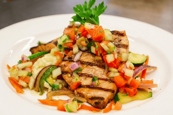 Grilled fish topped with fresh tomato and cucumber salsa, served over roasted vegetables on a white plate.