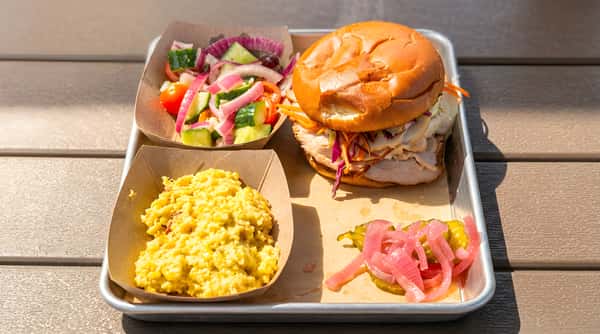 A turkey sandwich with coleslaw and carrots, a side of cucumber and red onion salad, corn salad, and pickles on a silver tray.