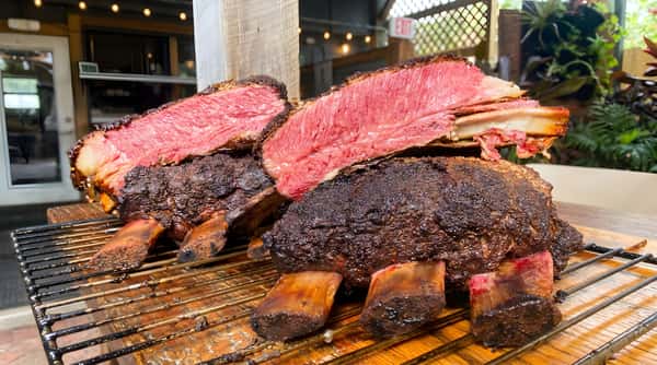Two large cuts of rare smoked prime rib with dark crust, resting on the bone on a grill rack.