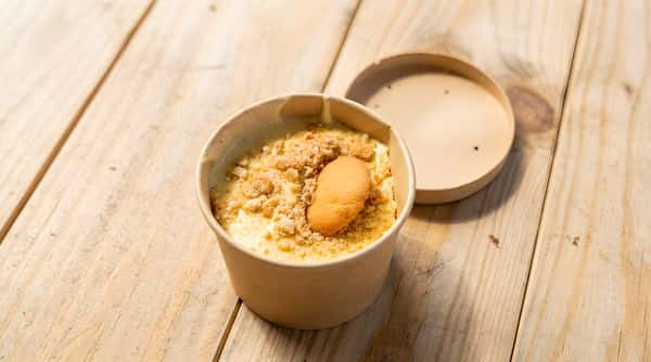 Banana pudding in a paper bowl with wafer crumbs and a whole vanilla wafer, on a wooden surface.