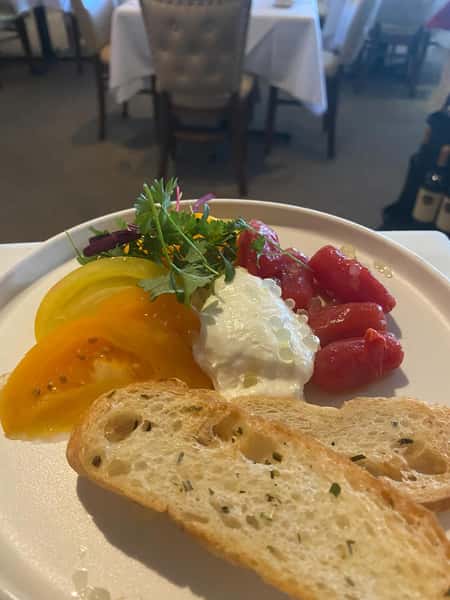 Heirloom tomato salad with burrata cheese, microgreens, and toasted baguette slices on a white plate.