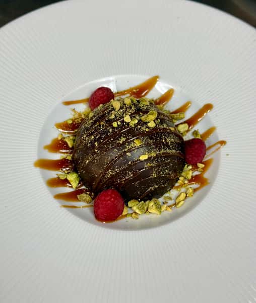 Chocolate dome drizzled with caramel sauce, garnished with crushed pistachios and fresh raspberries on a white plate.