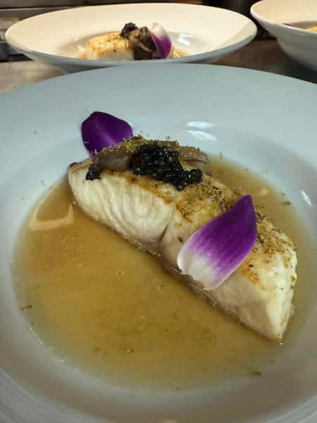 Poached white fish fillet served in a delicate broth, topped with black caviar, mushrooms, and garnished with crushed seasoning and vibrant flower petals.