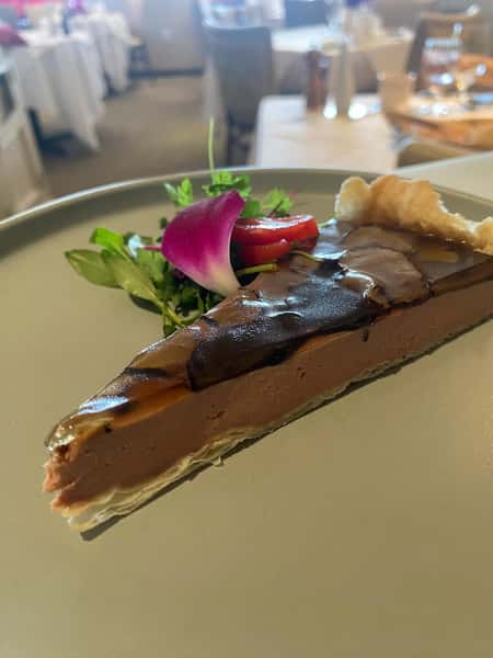 Slice of rich chocolate tart with a glossy top, served with microgreens, strawberries, and a pink flower petal garnish.