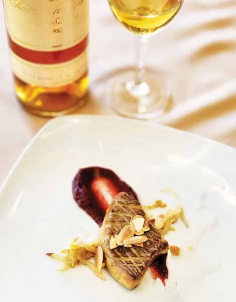 Seared foie gras garnished with toasted almonds, served over a red fruit reduction with wine and a bottle in the background.