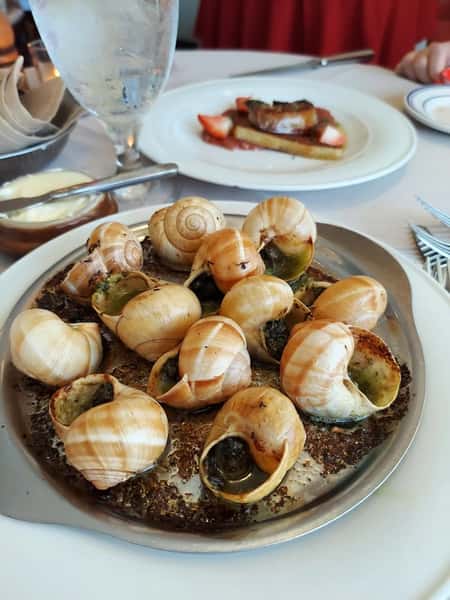 Baked escargot in garlic herb butter served in shells on a metal dish, with foie gras and strawberries in the background.
