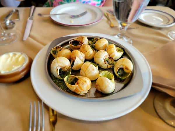 Escargot baked in garlic parsley butter, served in shells on a round metal dish over a white plate at a restaurant table.