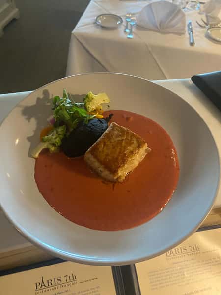 Seared fish fillet over creamy tomato sauce, served with roasted vegetables, a black potato, and fresh microgreens.