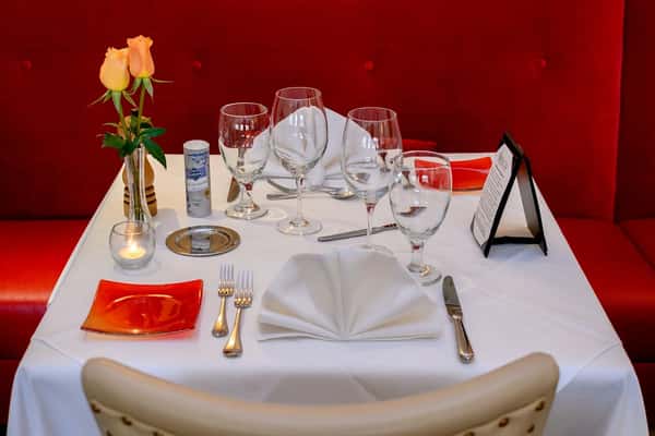 Fine dining table set with folded napkins, multiple glasses, two yellow roses in a vase, and red accent plates against red seating.