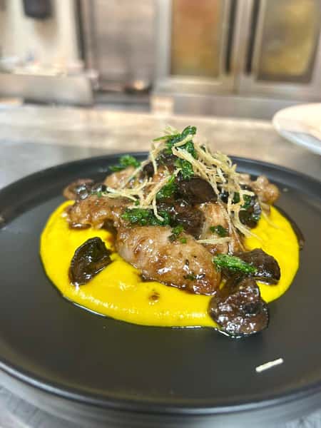 Veal sweetbreads on butternut squash purée with wild mushrooms, crispy leeks, and fried herbs on a black plate.