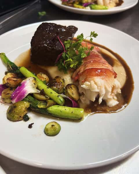 Elegant surf and turf dish with braised beef, lobster tail, mashed potatoes, asparagus, Brussels sprouts, and a rich brown sauce.