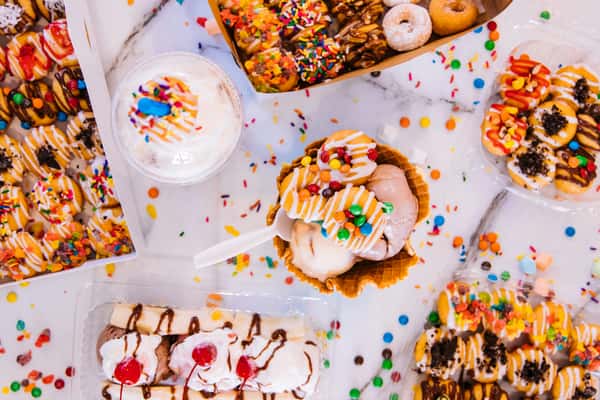 A variety of colorful donuts, ice cream in a cone, and a milkshake with whipped cream, all decorated with sprinkles and toppings on a marble surface.