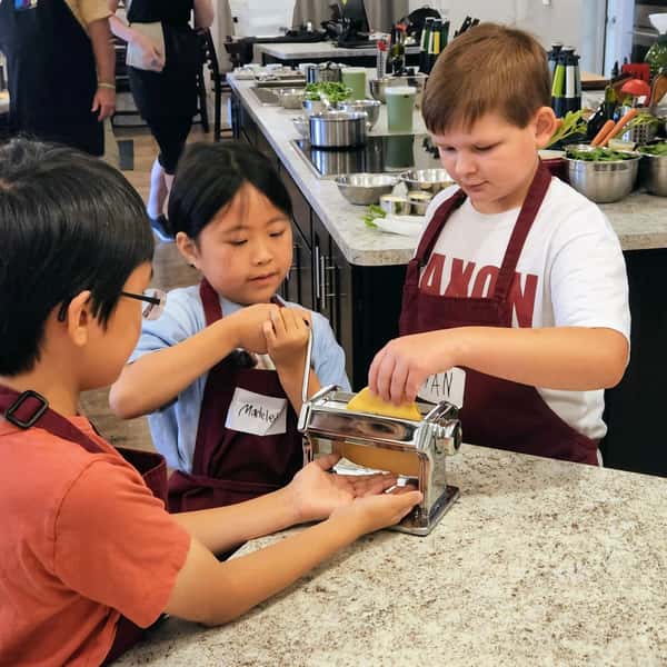 Children participating in a hands-on cooking class, using a pasta machine to roll fresh dough in a professional teaching kitchen.