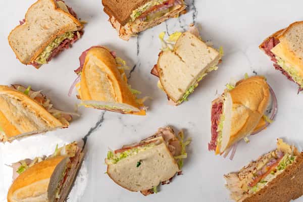 Assorted deli sandwiches with sliced meats, cheese, lettuce, and tomatoes on white and wheat bread, arranged on a marble surface in a casual restaurant setting