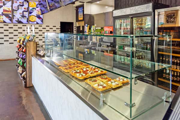 Modern pizza shop interior with glass display case showcasing fresh square slices, beverage cooler, and menu boards in a casual fast-casual restaurant setting