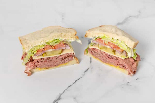 Pastrami sandwich halves with cheese, lettuce, tomato, pickles, and onions on soft bread, served on marble surface