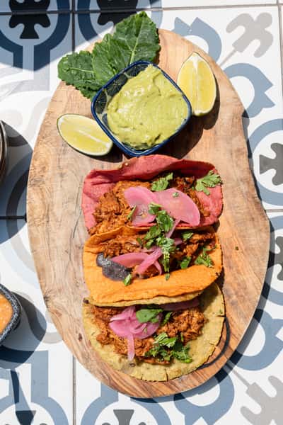 Trio of colorful street-style tacos with shredded meat, pickled red onions, and fresh cilantro on a wooden platter, served with guacamole and lime wedges.