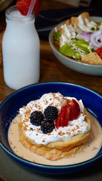 Decadent pastry soaked in sweet milk, topped with whipped cream, blackberries, and strawberries, served in a dessert bowl with a side of fresh strawberry horchata—perfect for showcasing indulgent Latin-inspired desserts.