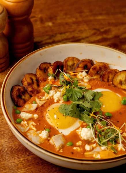 Savory breakfast stew featuring sunny-side-up eggs, tomato sauce, crumbled cheese, peas, and fried plantains, garnished with microgreens and fresh herbs.