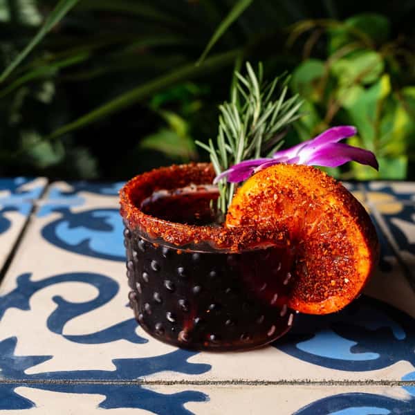Spicy mezcal cocktail served in a textured glass with a chili-lime rim, garnished with a dried orange slice, rosemary sprig, and edible orchid.