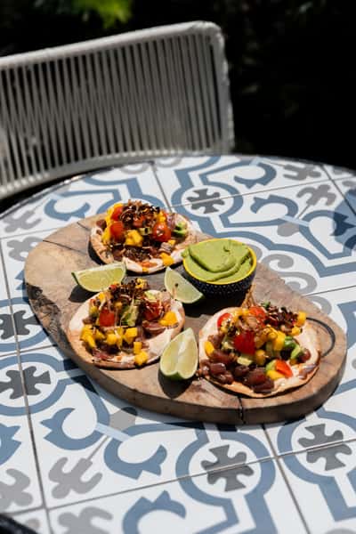 Trio of vibrant tuna tostadas topped with avocado, mango, tomato, and crispy onions, served with fresh lime wedges and creamy guacamole on a rustic board.