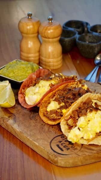 Breakfast tacos filled with scrambled eggs and crispy shredded beef, served on colorful corn tortillas with fresh guacamole and lime wedges—perfect for hearty Mexican breakfast menus and brunch offerings.