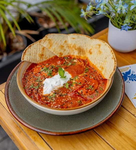 Hearty bowl of tomato-based curry garnished with fresh herbs, sesame seeds, and a dollop of cream, accompanied by soft pita bread.