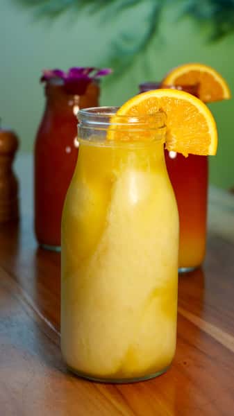 Refreshing orange juice served in a glass bottle with a slice of fresh orange garnish, set against a tropical background.