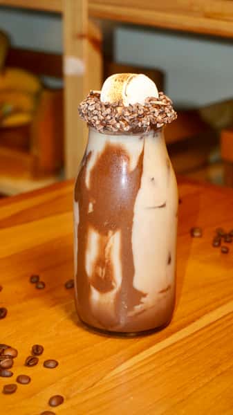 Iced mocha latte in a glass bottle with chocolate swirls and a toasted marshmallow topping, rimmed with chocolate bits and surrounded by coffee beans.