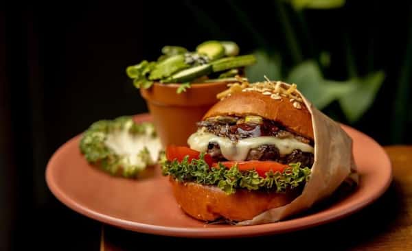 Gourmet cheeseburger with caramelized onions, lettuce, tomato, and melted cheese on a seeded brioche bun, served with a side salad in a terracotta cup.