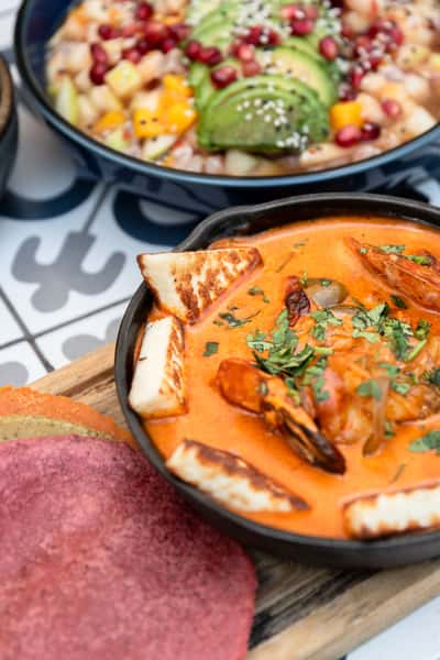 Rich shrimp and halloumi stew in a creamy orange sauce, garnished with fresh cilantro and served with vibrant tri-color corn tortillas; a standout dish in coastal Mexican cuisine with a fresh avocado and fruit ceviche bowl in the background.