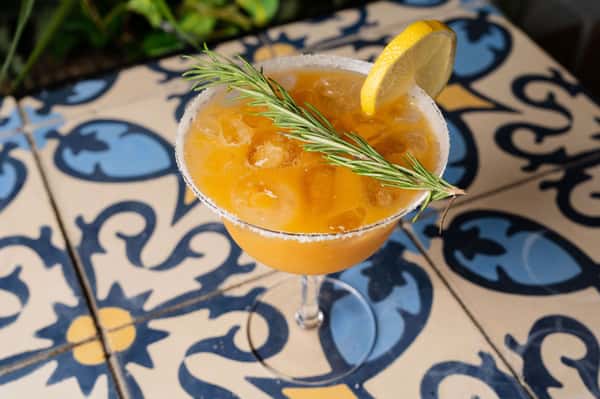 Iced orange cocktail served in a sugar-rimmed margarita glass, garnished with a lemon wheel and rosemary sprig on a colorful tile table.