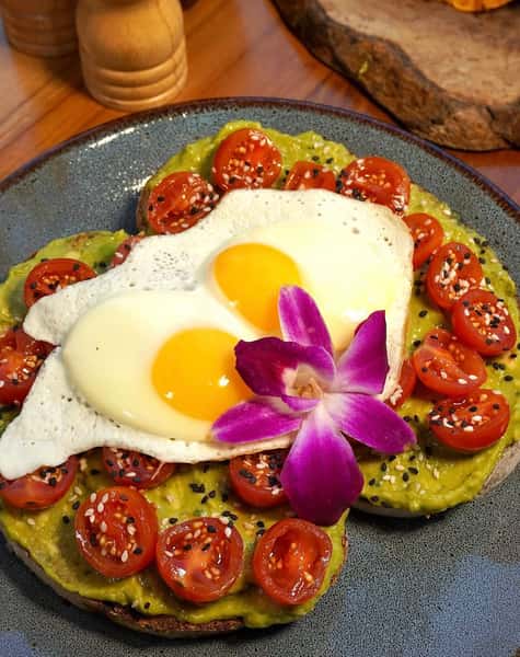 Vibrant avocado toast topped with cherry tomatoes, sunny-side-up eggs, and a purple orchid flower, sprinkled with sesame seeds for a gourmet brunch presentation.