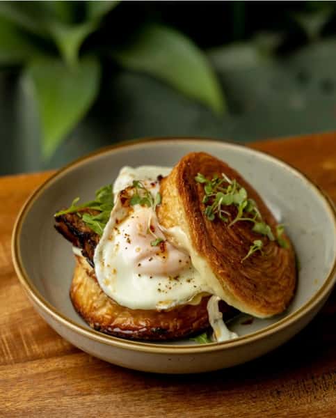 Gourmet breakfast sandwich with a fried egg, greens, and savory filling, served on a flaky croissant bun—perfect for elevated brunch menus and artisanal café bites.