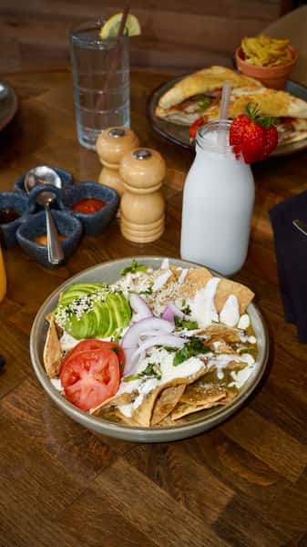 Traditional Mexican chilaquiles with green salsa, topped with avocado, tomato, red onion, crema, and queso fresco, served with a strawberry-topped milk beverage.