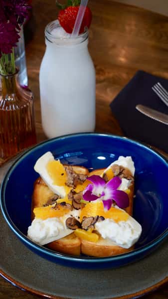 Brioche toast topped with whipped cream, fresh orange slices, and truffle shavings, garnished with an orchid flower, served with a chilled strawberry-topped milk beverage.
