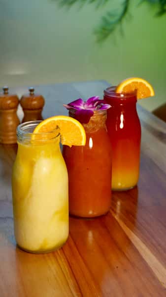 Trio of vibrant tropical juices served in glass bottles, garnished with orange slices and a purple orchid flower, featuring a yellow citrus blend, a red tropical mix, and a layered orange sunrise juice on a wooden table.