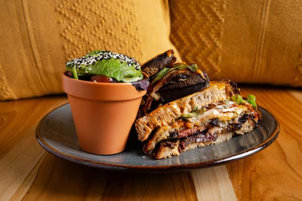 Smoky brisket melt sandwich layered with melted cheese and grilled vegetables on rustic sourdough bread, served with a side salad of avocado, cherry tomatoes, and sesame seeds in a terracotta-style cup.