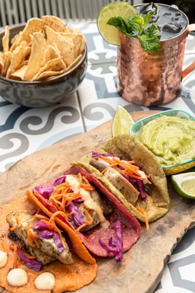 Colorful grilled fish tacos on orange, pink, and green tortillas, topped with cabbage, carrots, and aioli, served with guacamole, tortilla chips, and a refreshing cocktail.